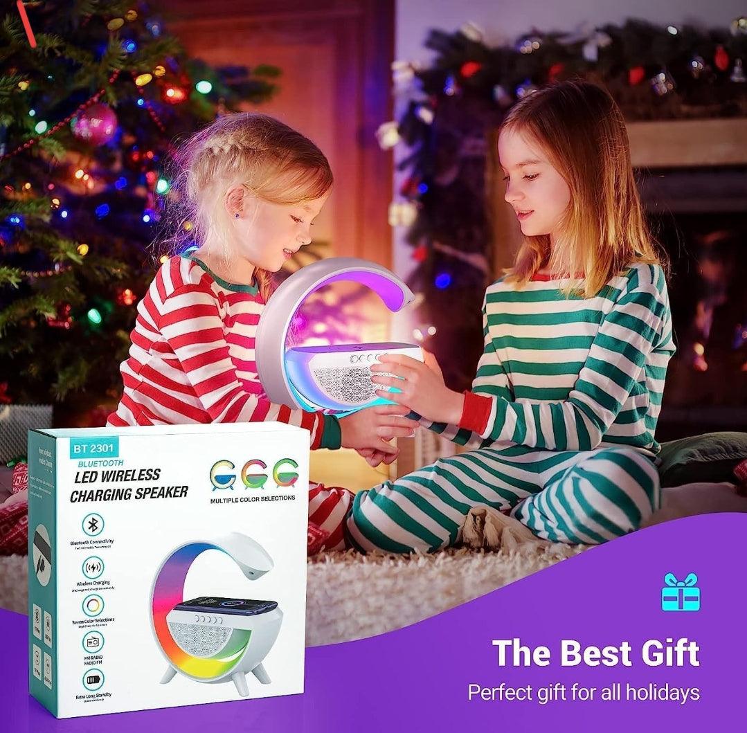 Two children in striped pajamas holding a G-shape LED table lamp with wireless charger and Bluetooth speaker near a Christmas tree, product box in foreground.