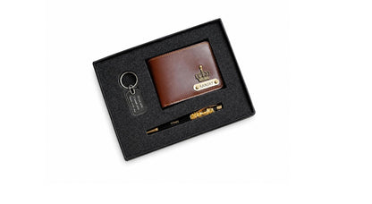 A luxury men's gift set in a black presentation box, featuring a brown leather wallet with a gold crown logo, a black pen with gold detailing, and a black keychain.